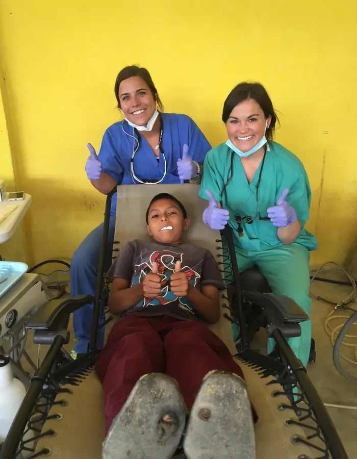 Two volunteers giving a thumbs up beside a boy in the dental chair, 2017.